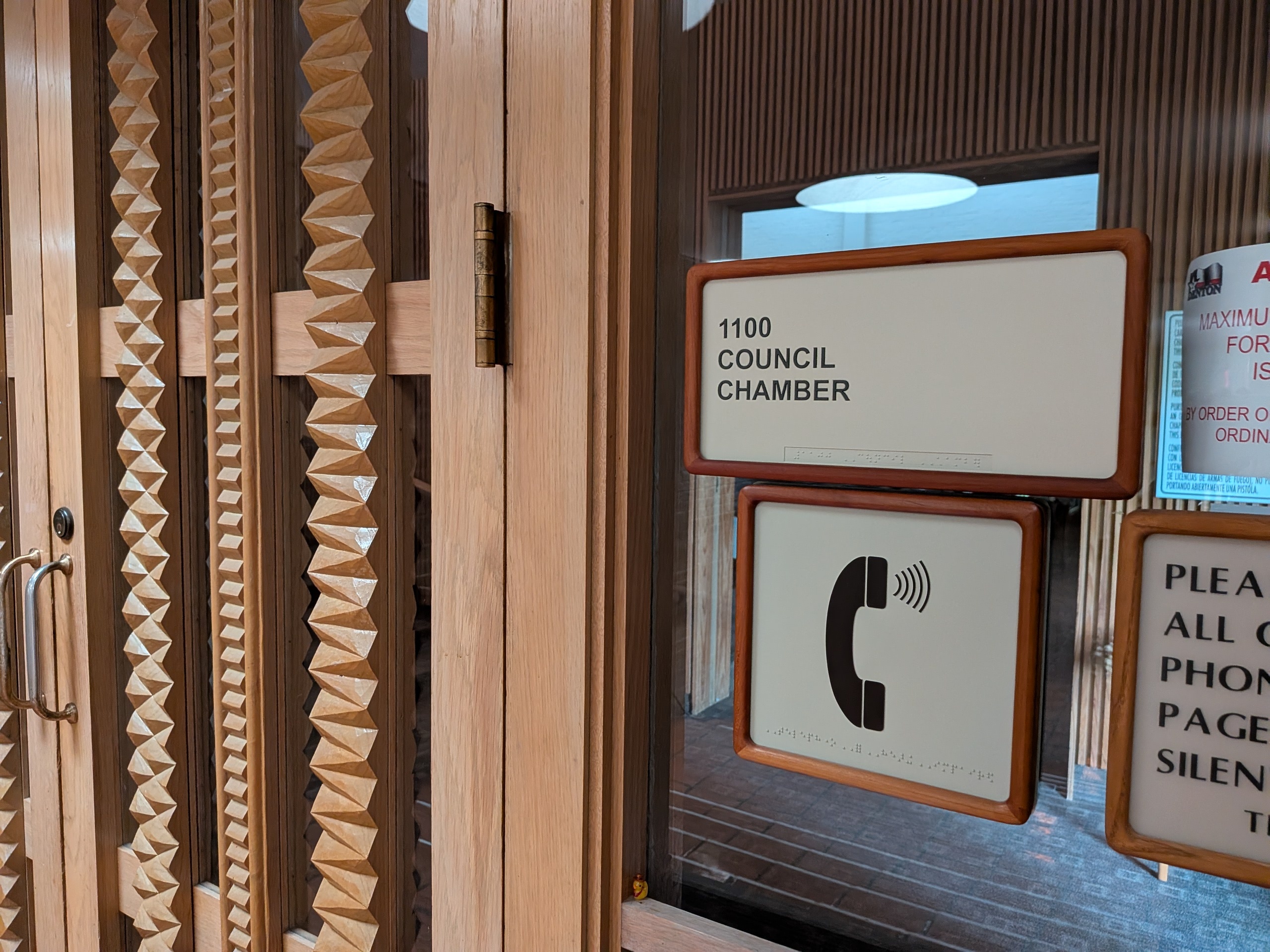 Photo of the signage outside the Denton City Hall Chambers.  The room number is 1100 Council Chamber.  There is an occupancy sign posted. Braille is below the corresponding text. 