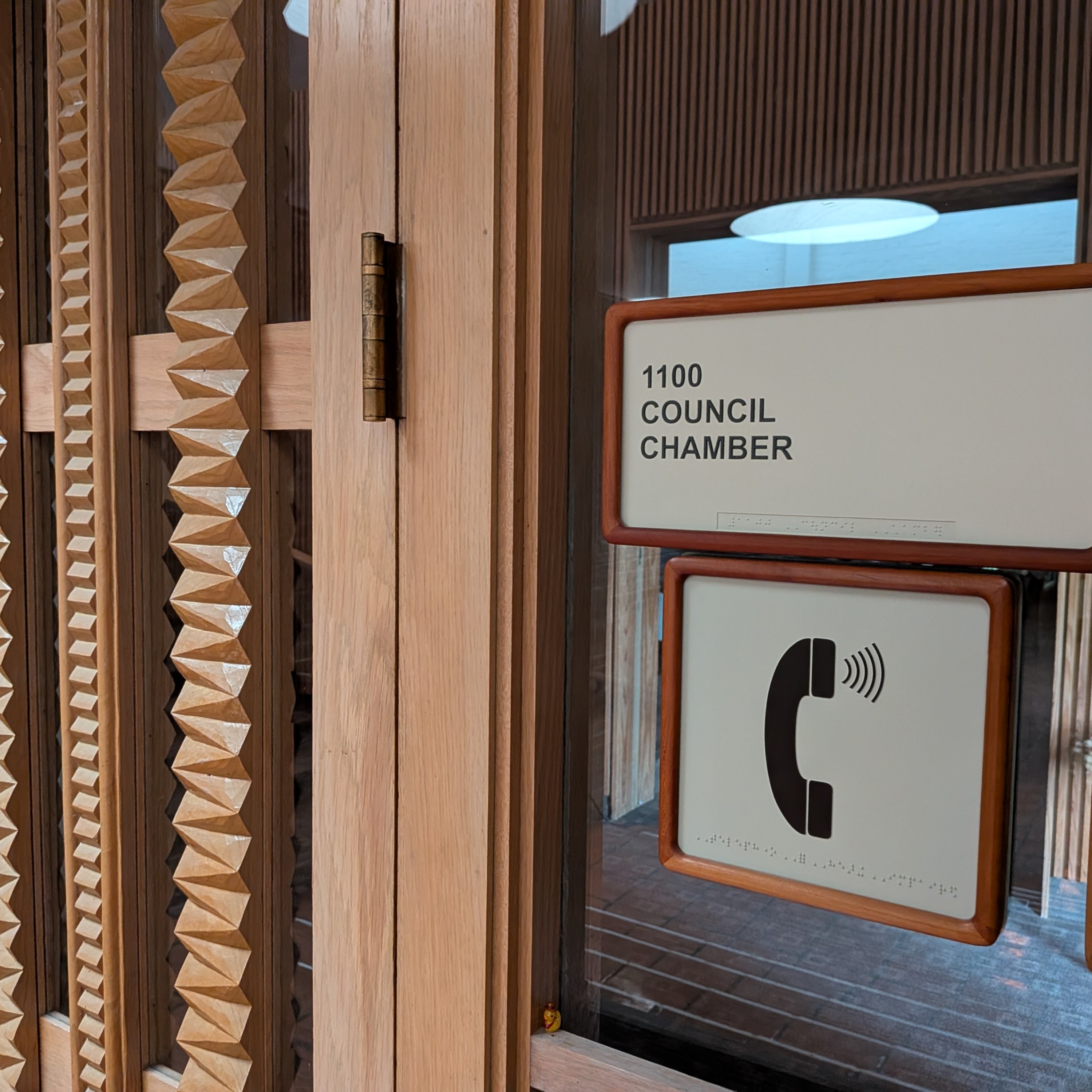 Photo of the signage outside the Denton City Hall Chambers.  The room number is 1100 Council Chamber.  There is an occupancy sign posted. Braille is below the corresponding text. 